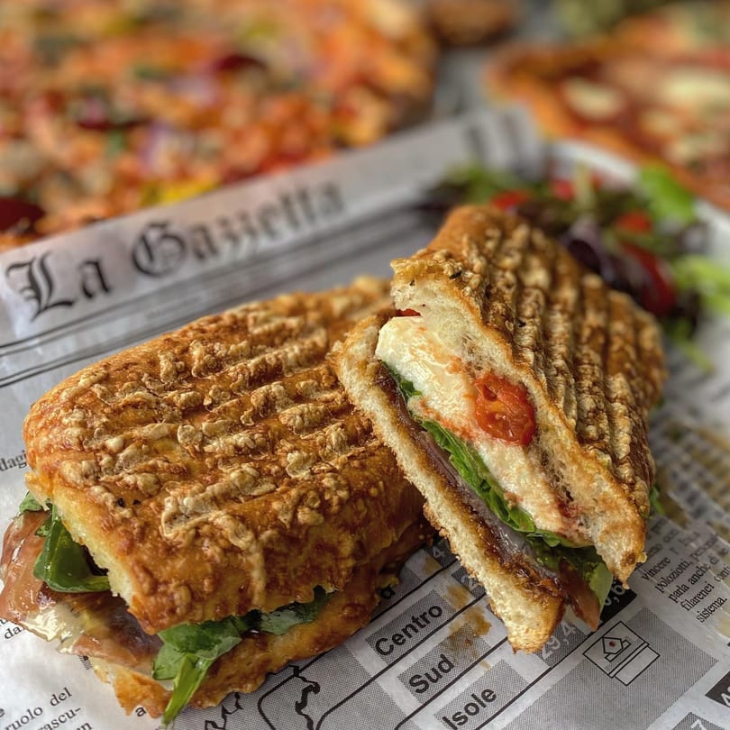 A grilled Italian panini, cut in half, filled with prosciutto, mozzarella, and greens, on newspaper.