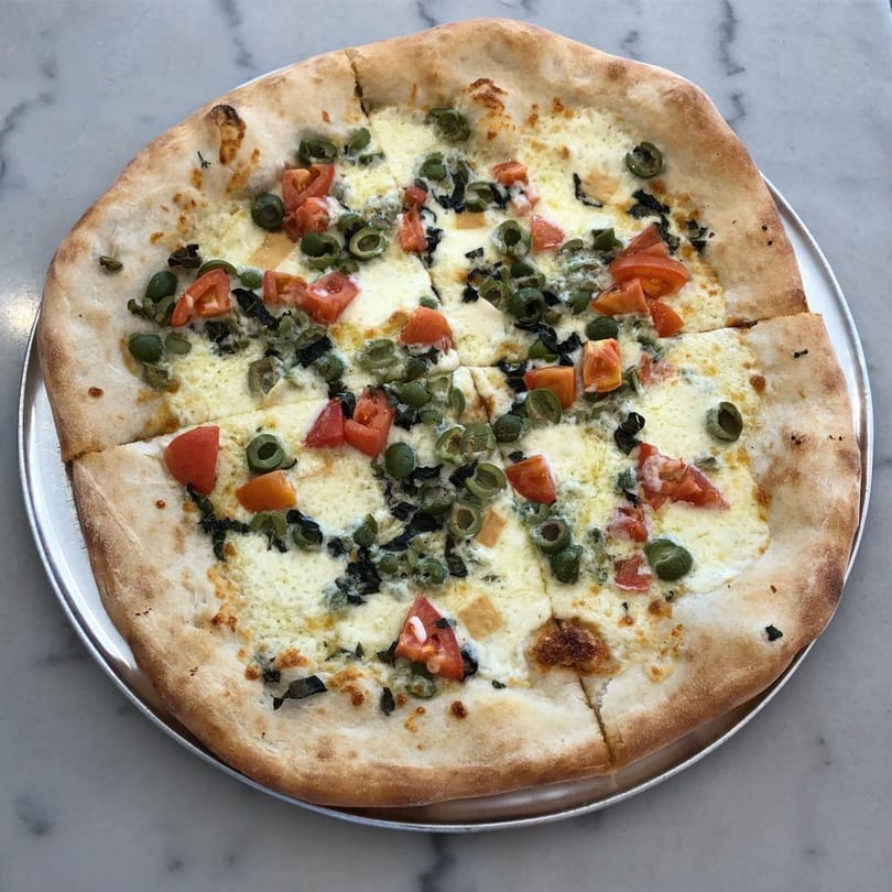 Becca's Kitchen Italian pizza with white cheese, green olives, diced tomatoes, and basil on a metal tray.