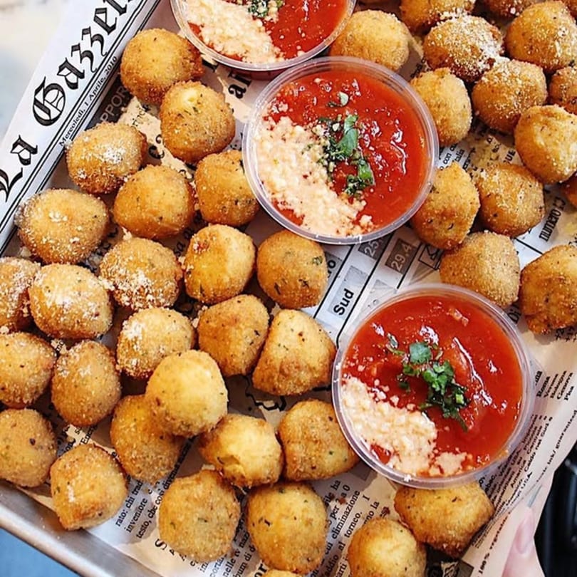 Golden-brown fried arancini balls with three cups of marinara sauce and grated cheese on newspaper.