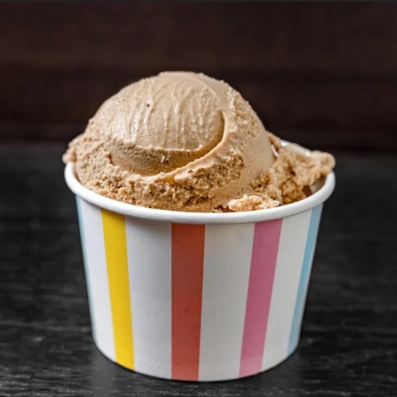 A scoop of light brown ice cream in a colorful striped paper cup on a dark surface.